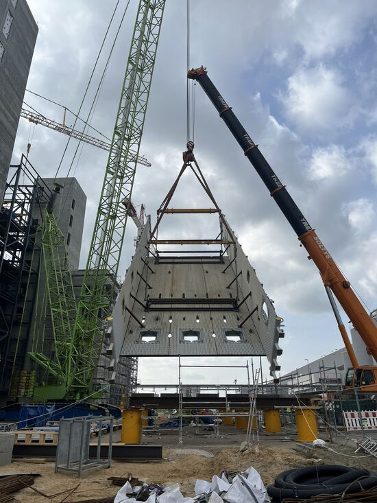 Ein großer Kran hebt ein massives Bauelement auf einer Baustelle in den Himmel, während Arbeiter die Konstruktion überwachen.