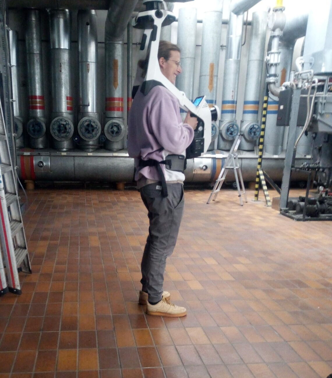Ein Mann in Arbeitskleidung trägt ein technisches Gerät in einem industriellen Raum mit großen Metallrohren.