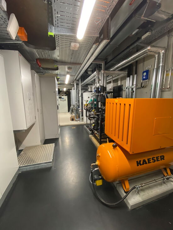 Ein moderner Technikraum mit einem orangefarbenen Kaeser-Kompressor und komplexer Verkabelung ist zu sehen.