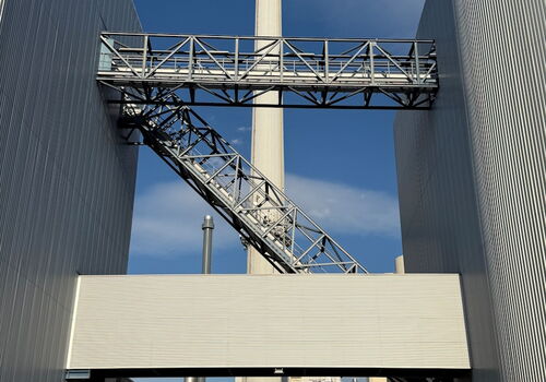 Ein hoher Schornstein erhebt sich zwischen zwei industriellen Gebäuden unter einem klaren blauen Himmel.
