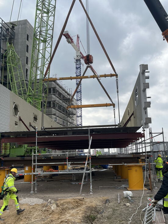 Bauarbeiter montieren mit großen Kränen ein Brückenelement auf einer Baustelle bei bewölktem Himmel.