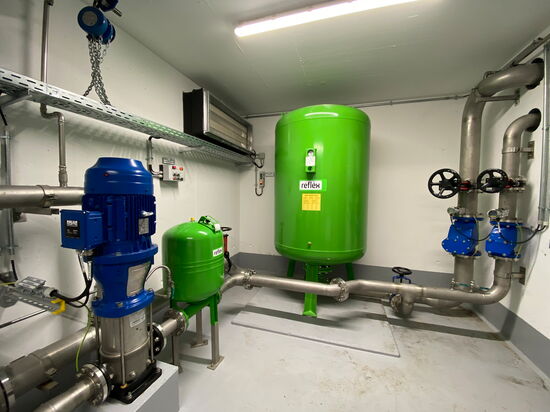 Ein technischer Raum zeigt eine grüne Druckausgleichsbehälteranlage mit blauen Pumpen und Rohrleitungen.