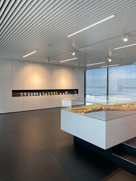 Ein moderner Ausstellungsraum mit einem Architekturmodell und farbenfrohen Broschüren auf einem Regal an der Wand.