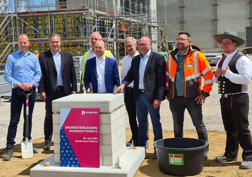 Eine Gruppe von Männern steht vor einem Grundstein mit der Aufschrift "Grundsteinlegung Progroup Power 2" auf einer Baustelle.