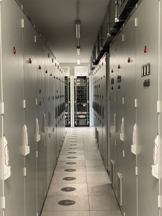 Ein langer, schmaler Flur in einem Serverraum ist gesäumt von grauen Schaltschränken und beleuchtet von Deckenlampen.