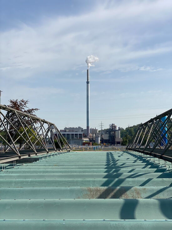 Eine Brücke führt zu einem Fabrikschornstein, aus dem weißer Rauch in den blauen Himmel aufsteigt.