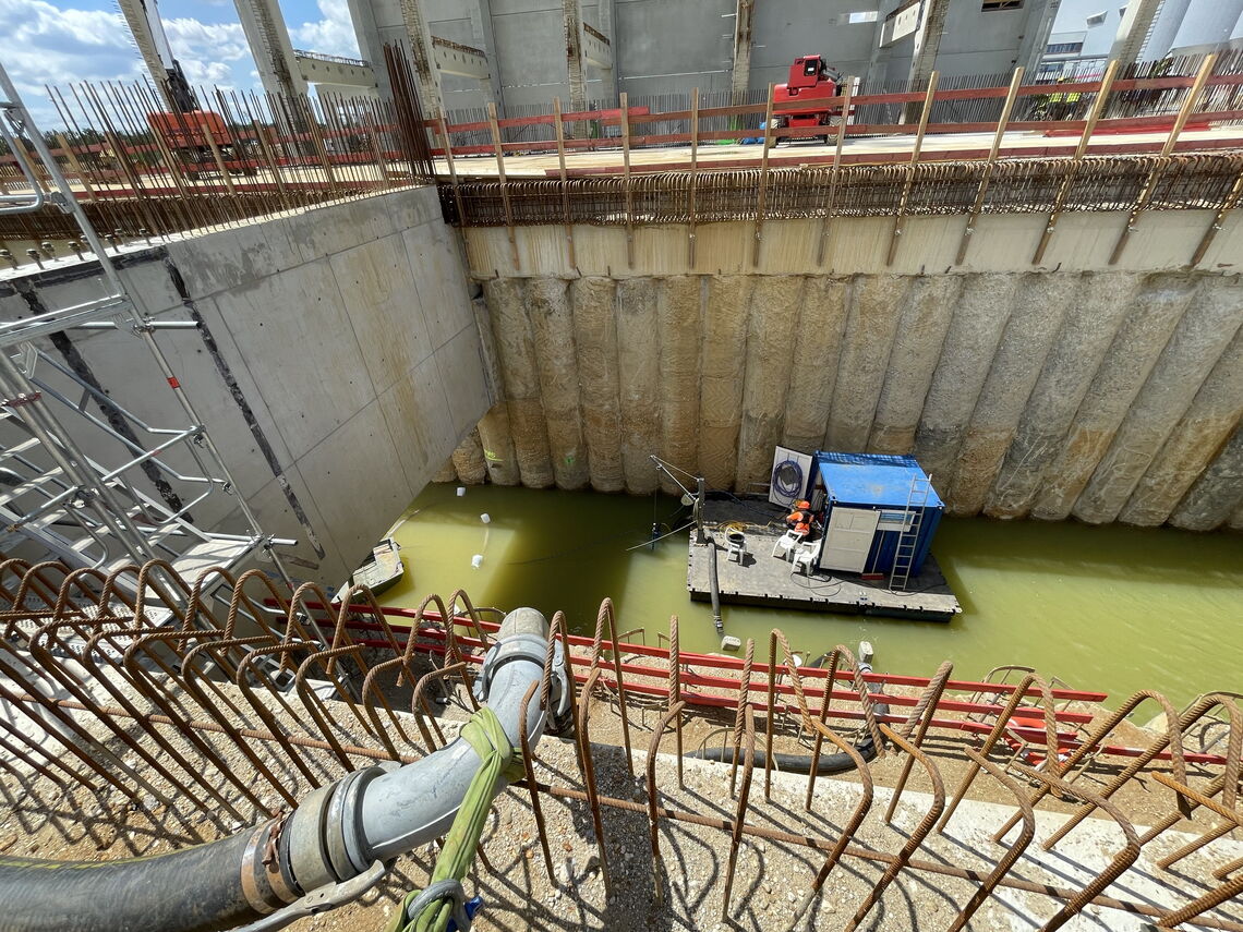 Ein Bauarbeiter sitzt in einer blauen Kabine auf einer Plattform über grünlichem Wasser, umgeben von einer Baustelle.