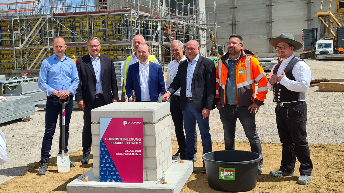 Eine Gruppe von Männern steht vor einem Grundstein mit der Aufschrift "Grundsteinlegung Progroup Power 2" auf einer Baustelle.