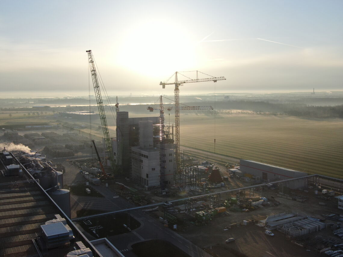 Eine Drohnenaufnahme zeigt eine Baustelle mit Kränen bei Sonnenaufgang, umgeben von weiten Feldern und Industrieanlagen.