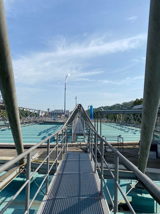 Ein langer Metallsteg führt über eine industrielle Anlage mit türkisfarbenen Wasserbecken unter klarem Himmel.