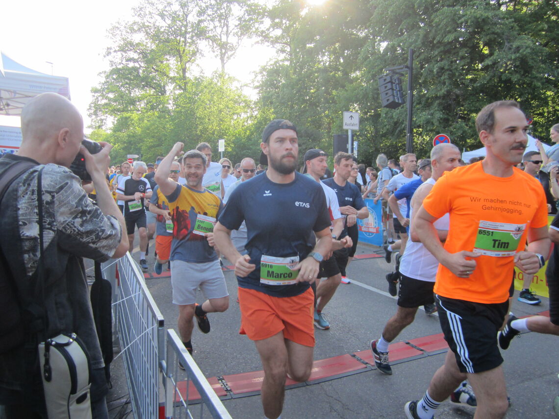 Läufer in bunten Trikots überqueren die Ziellinie bei einem sonnigen Straßenlauf, während ein Fotograf den Moment festhält.